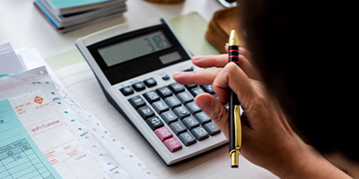 Mão de uma pessoa utilizando uma calculadora para tarefas financeiras, com documentos e contas espalhados sobre a mesa, simbolizando a precisão na gestão financeira e operacional proporcionada pelas soluções ERP da Alternativa Sistemas.